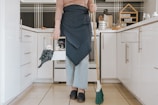 A person is standing in a modern kitchen holding a broom in one hand and a cleaning caddy in the other. They are wearing light-wash jeans, a brown long-sleeve shirt, and a dark apron. The kitchen features white cabinets, and the countertop has various kitchen items arranged neatly, including spices stored in a wooden rack.