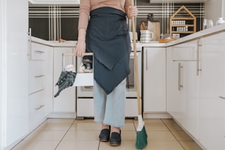 A person is standing in a modern kitchen holding a broom in one hand and a cleaning caddy in the other. They are wearing light-wash jeans, a brown long-sleeve shirt, and a dark apron. The kitchen features white cabinets, and the countertop has various kitchen items arranged neatly, including spices stored in a wooden rack.