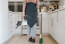 A person is standing in a modern kitchen holding a broom in one hand and a cleaning caddy in the other. They are wearing light-wash jeans, a brown long-sleeve shirt, and a dark apron. The kitchen features white cabinets, and the countertop has various kitchen items arranged neatly, including spices stored in a wooden rack.