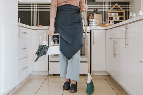 A person is standing in a modern kitchen holding a broom in one hand and a cleaning caddy in the other. They are wearing light-wash jeans, a brown long-sleeve shirt, and a dark apron. The kitchen features white cabinets, and the countertop has various kitchen items arranged neatly, including spices stored in a wooden rack.
