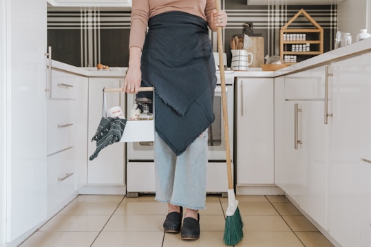 A person is standing in a modern kitchen holding a broom in one hand and a cleaning caddy in the other. They are wearing light-wash jeans, a brown long-sleeve shirt, and a dark apron. The kitchen features white cabinets, and the countertop has various kitchen items arranged neatly, including spices stored in a wooden rack.