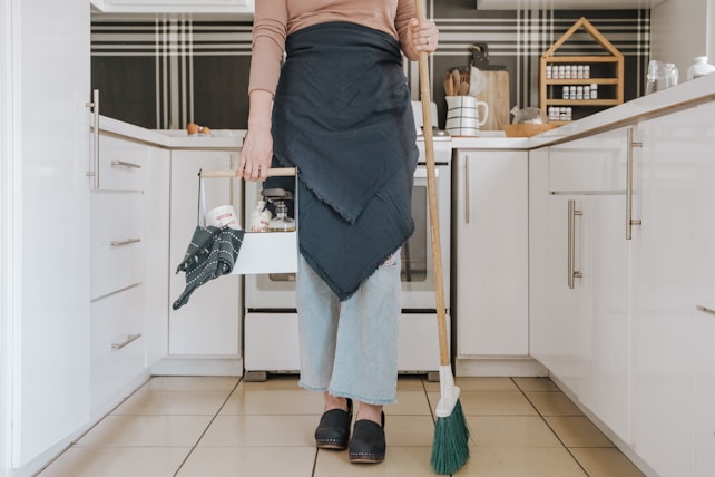 A person is standing in a modern kitchen holding a broom in one hand and a cleaning caddy in the other. They are wearing light-wash jeans, a brown long-sleeve shirt, and a dark apron. The kitchen features white cabinets, and the countertop has various kitchen items arranged neatly, including spices stored in a wooden rack.