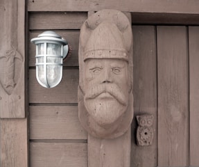 A wooden wall features a rustic outdoor light fixture next to a carved wooden figure of a bearded man wearing a helmet. The wood displays a weathered texture, and there is an additional small owl carving below the face.