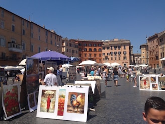 A bustling outdoor art market set against a backdrop of historic buildings. Numerous art pieces, including paintings of landscapes and animals, are displayed on easels. People are strolling through the market, some browsing the artworks under the shade of umbrellas. The sky is clear and blue, adding to the vibrant atmosphere of the scene.