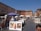A bustling outdoor art market set against a backdrop of historic buildings. Numerous art pieces, including paintings of landscapes and animals, are displayed on easels. People are strolling through the market, some browsing the artworks under the shade of umbrellas. The sky is clear and blue, adding to the vibrant atmosphere of the scene.