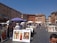 A bustling outdoor art market set against a backdrop of historic buildings. Numerous art pieces, including paintings of landscapes and animals, are displayed on easels. People are strolling through the market, some browsing the artworks under the shade of umbrellas. The sky is clear and blue, adding to the vibrant atmosphere of the scene.
