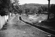 A winding dirt path stretches alongside a construction area. The foreground features a narrow, unpaved road bordered by a low concrete wall on one side and a trench on the other. Sparse trees and scattered debris can be seen along the pathway. In the distance, hills and a dense cluster of trees are visible, suggesting a rural or semi-urban setting.