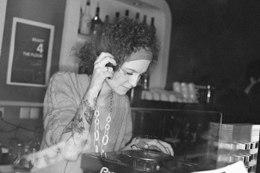 A person with curly hair and a headband, wearing a sweater and a chain necklace, operates DJ equipment in a dimly lit room. The person's arm is adorned with tattoos, and they appear focused on the task. Behind them, there are shelves with bottles and other items.