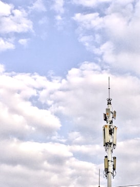 A telecommunications tower stands against a backdrop of a partly cloudy sky. The structure is equipped with various antennas and equipment, suggesting its use for communication purposes.