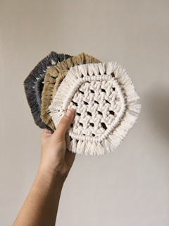 A hand holding three woven coasters with fringe edges. The coasters appear to be made from natural fibers, displaying a circular pattern with intricate weaving. The colors transition from light beige to darker shades.