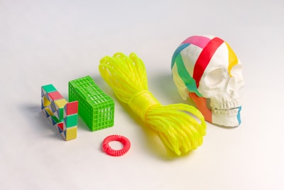 A collection of colorful objects is arranged on a light surface. The objects include a multicolored human skull with bright geometric-patterned sections, a yellow coiled rope, a green rectangular grid-like structure, a colorful geometric puzzle toy, and a small circular red coil.