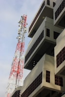 A tall, red and white telecommunication tower stands beside a modern building with geometric patterns and balconies. The building has a beige facade with brown lattice brickwork. The sky is clear with some clouds visible.
