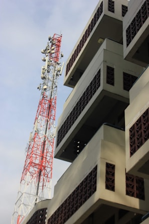A tall, red and white telecommunication tower stands beside a modern building with geometric patterns and balconies. The building has a beige facade with brown lattice brickwork. The sky is clear with some clouds visible.