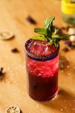 A tall glass filled with a deep red liquid is garnished with fresh mint leaves. The rim of the glass is coated with a chili salt mixture. Ice cubes are visible within the drink, and the glass is placed on a wooden surface scattered with dried lime slices and coarse salt.