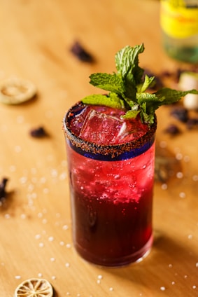 A tall glass filled with a deep red liquid is garnished with fresh mint leaves. The rim of the glass is coated with a chili salt mixture. Ice cubes are visible within the drink, and the glass is placed on a wooden surface scattered with dried lime slices and coarse salt.