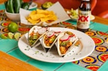 A white plate with three tacos filled with vegetables, broccoli, radishes, and salsa is arranged on a colorful, patterned tablecloth. In the background, a bowl of tortilla chips, sliced limes, jalapeños, guacamole, and a bottle of Dos Equis beer are visible.