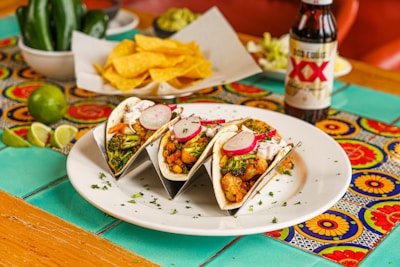 A white plate with three tacos filled with vegetables, broccoli, radishes, and salsa is arranged on a colorful, patterned tablecloth. In the background, a bowl of tortilla chips, sliced limes, jalapeños, guacamole, and a bottle of Dos Equis beer are visible.