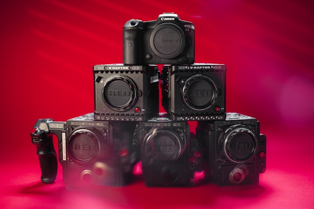 A collection of professional cameras is arranged against a vibrant red background. The setup includes multiple RED brand cameras and one Canon EOS R5 camera positioned at the top. The cameras are stacked in a pyramid-like fashion, emphasizing their robust and advanced design.