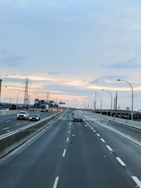 A wide highway with multiple lanes in both directions, featuring vehicles such as cars and a truck. The highway is bordered by concrete barriers and streetlights. In the background, there's an industrial setting with power lines and cranes. The sky is overcast with a hint of sunlight at the horizon.