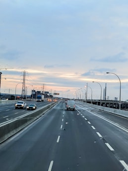 A wide highway with multiple lanes in both directions, featuring vehicles such as cars and a truck. The highway is bordered by concrete barriers and streetlights. In the background, there's an industrial setting with power lines and cranes. The sky is overcast with a hint of sunlight at the horizon.