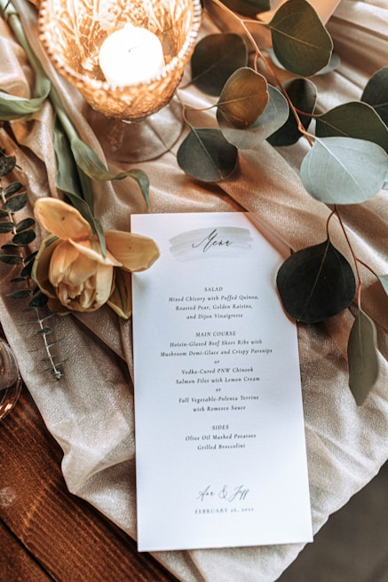 A wedding menu card placed on a textured fabric, surrounded by assorted greenery and eucalyptus leaves. A lit, intricately designed candle in a holder adds a warm glow to the scene, accompanied by delicate yellow flowers.