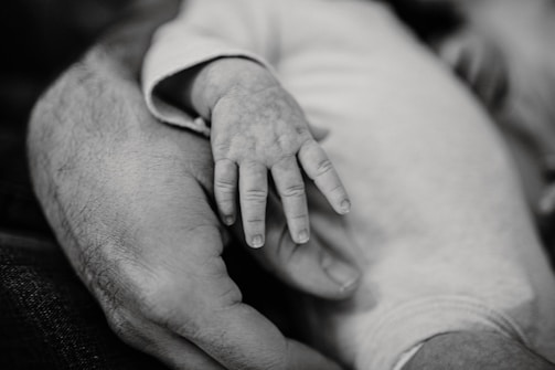 A black and white close-up of an adult hand gently holding a baby's hand. The baby's tiny fingers rest lightly on the adult's hand, depicting a tender moment of connection and warmth.
