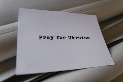 A piece of white paper with the words 'Pray for Ukraine' typed in a bold, black font is placed on a soft, light-colored fabric background.