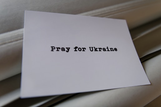 A piece of white paper with the words 'Pray for Ukraine' typed in a bold, black font is placed on a soft, light-colored fabric background.