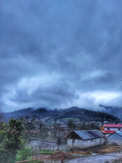 A rural landscape featuring a collection of small houses and a larger structure with a metal roof in the foreground. The background consists of rolling hills partially obscured by thick, dark clouds, suggesting an overcast or stormy day. Some vegetation, including a tree with green leaves, is visible at the edge of the frame.