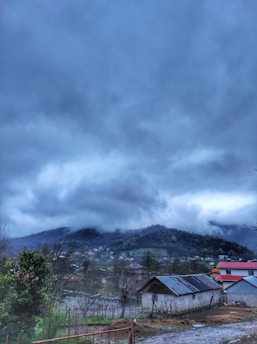 A rural landscape featuring a collection of small houses and a larger structure with a metal roof in the foreground. The background consists of rolling hills partially obscured by thick, dark clouds, suggesting an overcast or stormy day. Some vegetation, including a tree with green leaves, is visible at the edge of the frame.