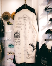 A white coat with hand-drawn black illustrations and text hangs on display. The artwork includes various sketches, including a masked face, glasses, lightning bolts, and statements like 'There’s no future for you' and 'New York.' It is surrounded by other clothing items and accessories, such as hats and sneakers, in a retail setting.