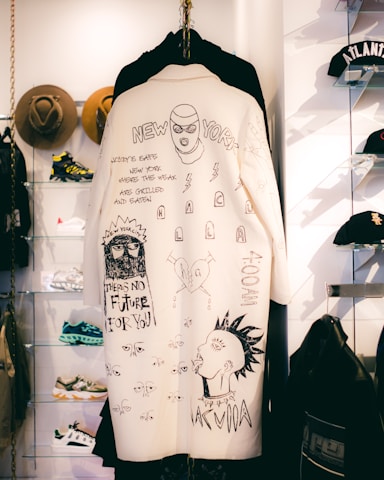 A white coat with hand-drawn black illustrations and text hangs on display. The artwork includes various sketches, including a masked face, glasses, lightning bolts, and statements like 'There’s no future for you' and 'New York.' It is surrounded by other clothing items and accessories, such as hats and sneakers, in a retail setting.