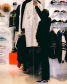 A person is selecting a white coat decorated with artistic drawings in a clothing store. The setting includes various apparel items such as hats, shoes, and jackets displayed on shelves and racks. The store has a modern ambiance with bright lighting.