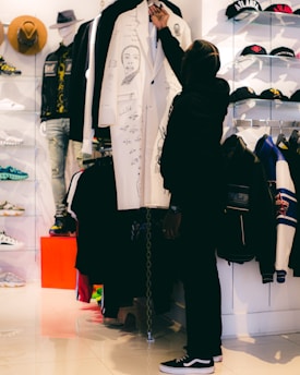 A person is selecting a white coat decorated with artistic drawings in a clothing store. The setting includes various apparel items such as hats, shoes, and jackets displayed on shelves and racks. The store has a modern ambiance with bright lighting.