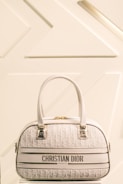 A luxury Christian Dior handbag with a cream and white color scheme is prominently displayed against a minimalist background featuring geometric patterns. The bag has two leather handles, gold hardware, and a textured surface with a repeating Dior logo pattern.