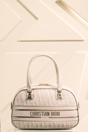 A luxury Christian Dior handbag with a cream and white color scheme is prominently displayed against a minimalist background featuring geometric patterns. The bag has two leather handles, gold hardware, and a textured surface with a repeating Dior logo pattern.