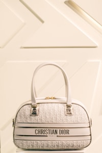 A luxury Christian Dior handbag with a cream and white color scheme is prominently displayed against a minimalist background featuring geometric patterns. The bag has two leather handles, gold hardware, and a textured surface with a repeating Dior logo pattern.