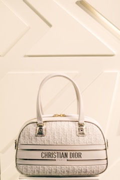 A luxury Christian Dior handbag with a cream and white color scheme is prominently displayed against a minimalist background featuring geometric patterns. The bag has two leather handles, gold hardware, and a textured surface with a repeating Dior logo pattern.