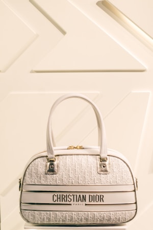 A luxury Christian Dior handbag with a cream and white color scheme is prominently displayed against a minimalist background featuring geometric patterns. The bag has two leather handles, gold hardware, and a textured surface with a repeating Dior logo pattern.