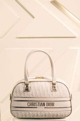 A luxury Christian Dior handbag with a cream and white color scheme is prominently displayed against a minimalist background featuring geometric patterns. The bag has two leather handles, gold hardware, and a textured surface with a repeating Dior logo pattern.