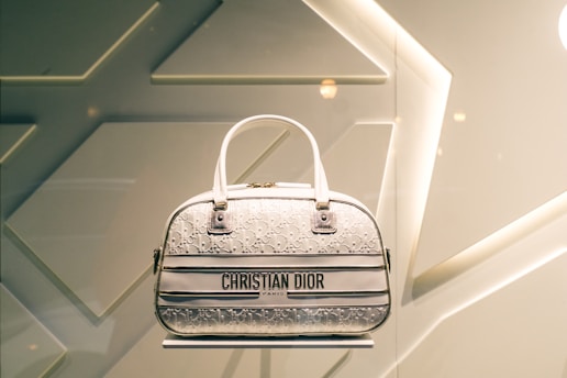 A designer handbag is displayed against a stylish, geometric patterned background. The bag features a light color palette with elegant design elements and prominently showcases the brand 'Christian Dior' on the front.