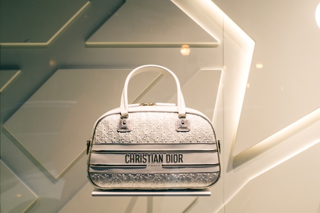 A designer handbag is displayed against a stylish, geometric patterned background. The bag features a light color palette with elegant design elements and prominently showcases the brand 'Christian Dior' on the front.