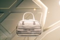 A designer handbag is displayed against a stylish, geometric patterned background. The bag features a light color palette with elegant design elements and prominently showcases the brand 'Christian Dior' on the front.