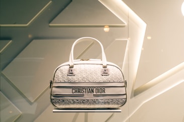A designer handbag is displayed against a stylish, geometric patterned background. The bag features a light color palette with elegant design elements and prominently showcases the brand 'Christian Dior' on the front.