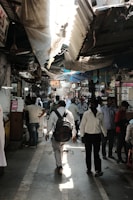 A crowded marketplace corridor with people walking in various directions. Overhead, there are makeshift tarps and damaged roofs providing partial shade. Vendors with small stalls line the sides, with stacked goods and signage. The atmosphere appears bustling and busy.