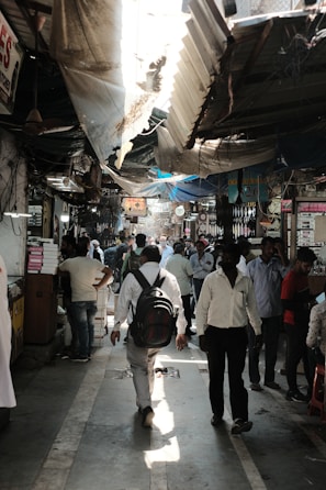 A crowded marketplace corridor with people walking in various directions. Overhead, there are makeshift tarps and damaged roofs providing partial shade. Vendors with small stalls line the sides, with stacked goods and signage. The atmosphere appears bustling and busy.