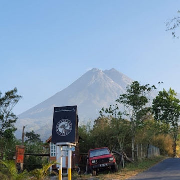 A mountain stands prominently in the background, partially covered with lush greenery. In the foreground, there is a small structure with a sign displaying a logo. A red vehicle is parked near the building amidst various trees and plants. The sky is clear, suggesting a calm day.