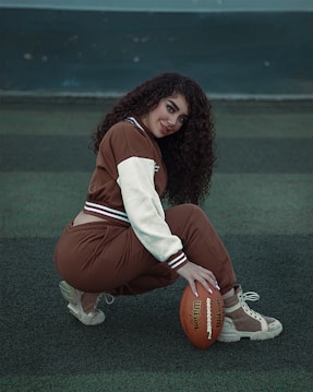 A person crouches on a sports field, wearing a brown and white jacket with matching pants and cream-colored sneakers. They are holding an American football, gazing toward the camera with a slight smile. The background consists of a dark green field with a net in the distance.