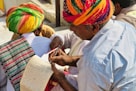 A group of people wearing colorful traditional attire, with one person in a vibrant turban writing or reviewing a paper using a pen. Another person also dressed in traditional clothing holds a notepad, which appears to have handwritten notes. The setting is outdoors with natural lighting.