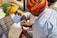 A group of people wearing colorful traditional attire, with one person in a vibrant turban writing or reviewing a paper using a pen. Another person also dressed in traditional clothing holds a notepad, which appears to have handwritten notes. The setting is outdoors with natural lighting.